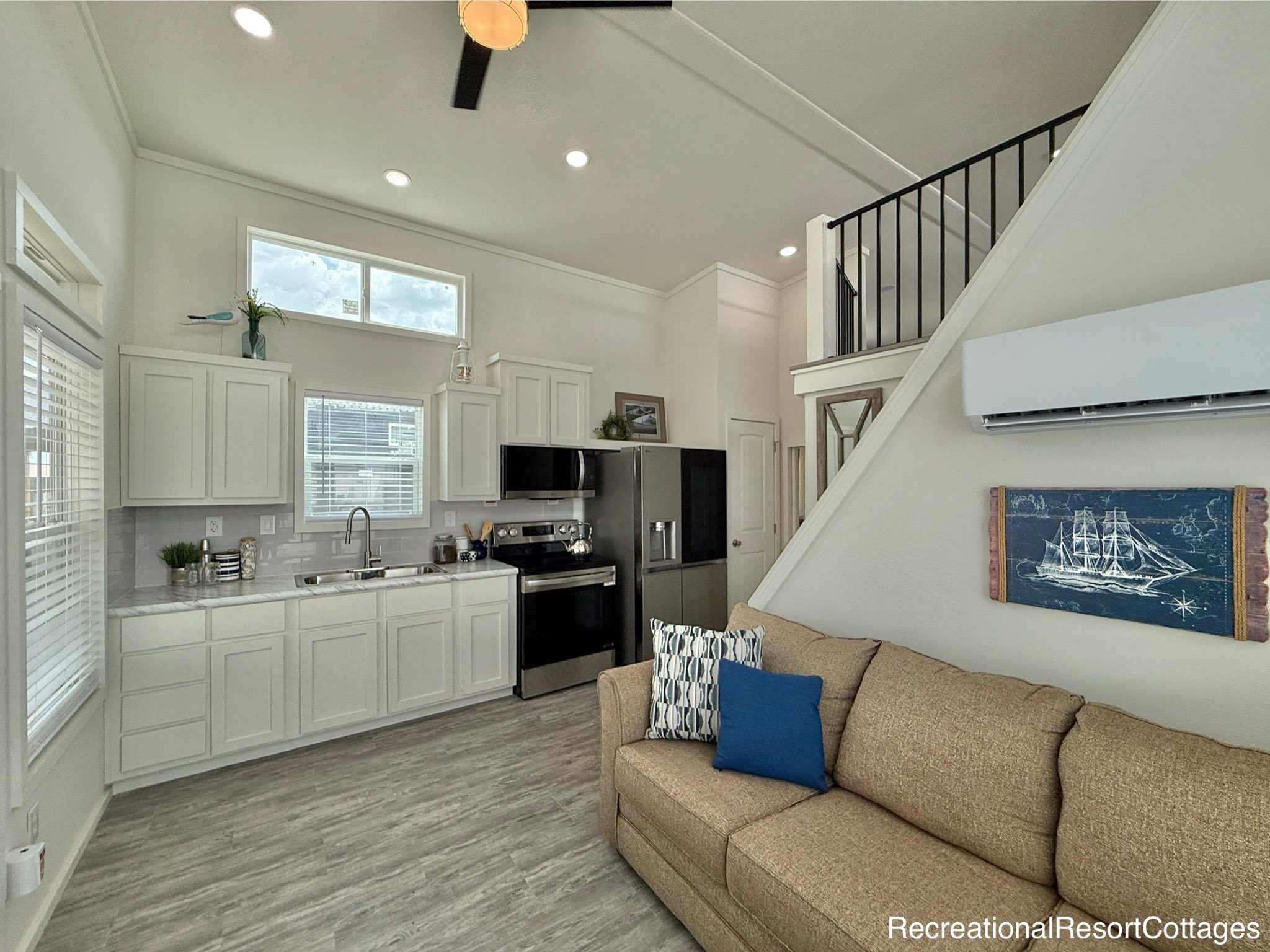 Modern tiny home interior with open kitchen and living area. White cabinets, stainless appliances, brown sofa, nautical artwork, and loft stairs. Cozy and bright.