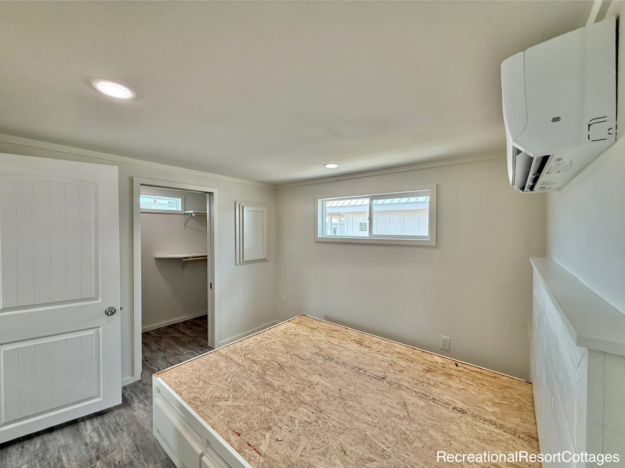 A minimalistic bedroom features a raised wooden bed frame, white walls, and small horizontal window. An open doorway reveals a closet with shelving.