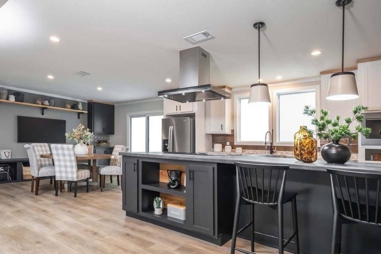 Modern kitchen with a large island and black chairs, pendant lighting, white cabinets, and stainless steel appliances. Dining area with plaid chairs.