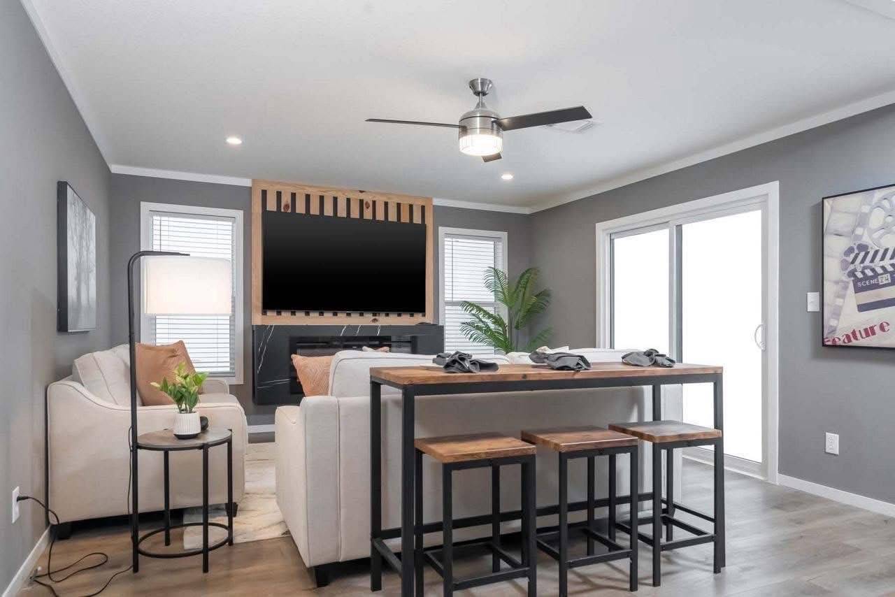 Modern living room with gray walls, a cozy white sofa, and a large TV. Wooden table with stools, ceiling fan, and tall plant add comfort and style.