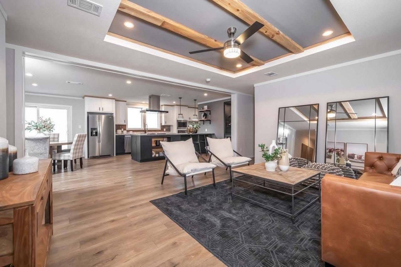Spacious modern living room; features a wooden ceiling accent, large mirrors, neutral tones, a brown leather sofa, and an open kitchen in the background.