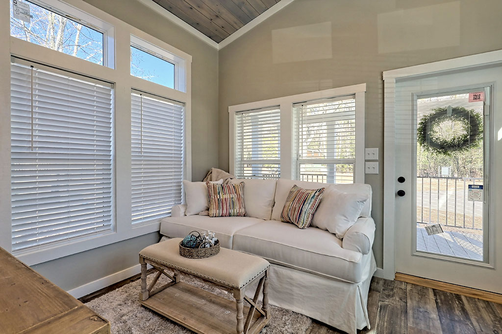 Cozy living room with a white sofa, striped pillows, and a beige coffee table. Sunlit from large windows with blinds. Door with a wreath leads outside.