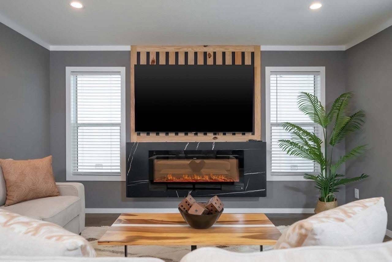 A modern living room with a wall-mounted TV above a sleek, marble electric fireplace. Beige couches, a wooden coffee table, and a potted plant add warmth.