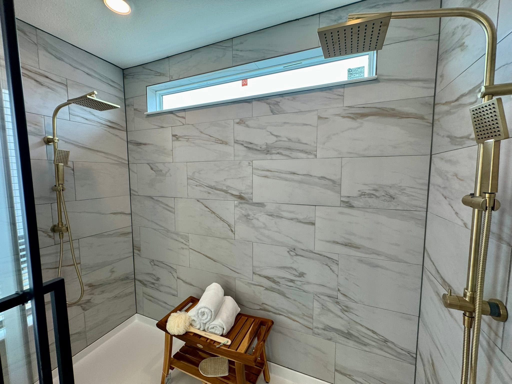 Luxurious shower with marble tiles, dual rainfall showerheads, and a wooden stool holding neatly rolled white towels, under soft ceiling lighting.