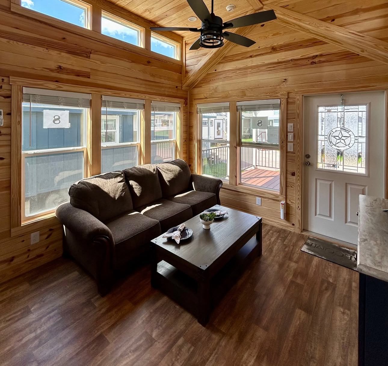 Cozy living room with wooden walls, large windows, and natural light. Features a dark sofa, coffee table, ceiling fan, and a decorative door.