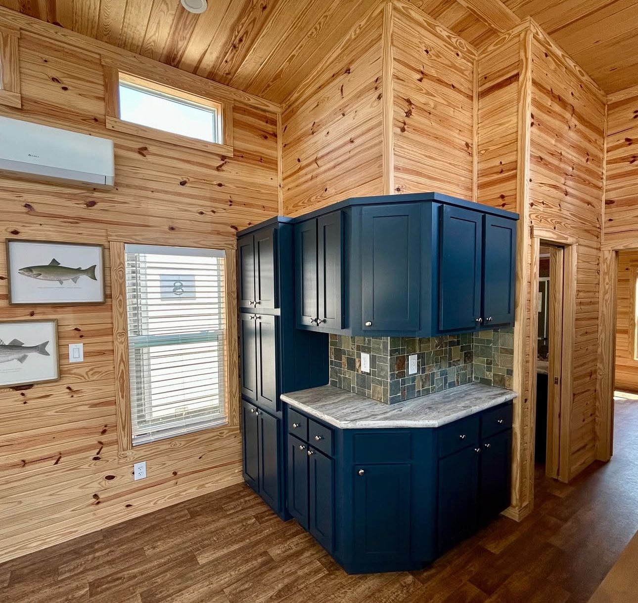 Cozy wooden interior with rich, knotty pine walls and ceiling. Dark blue kitchen cabinets and stone backsplash contrast, enhancing rustic warmth.