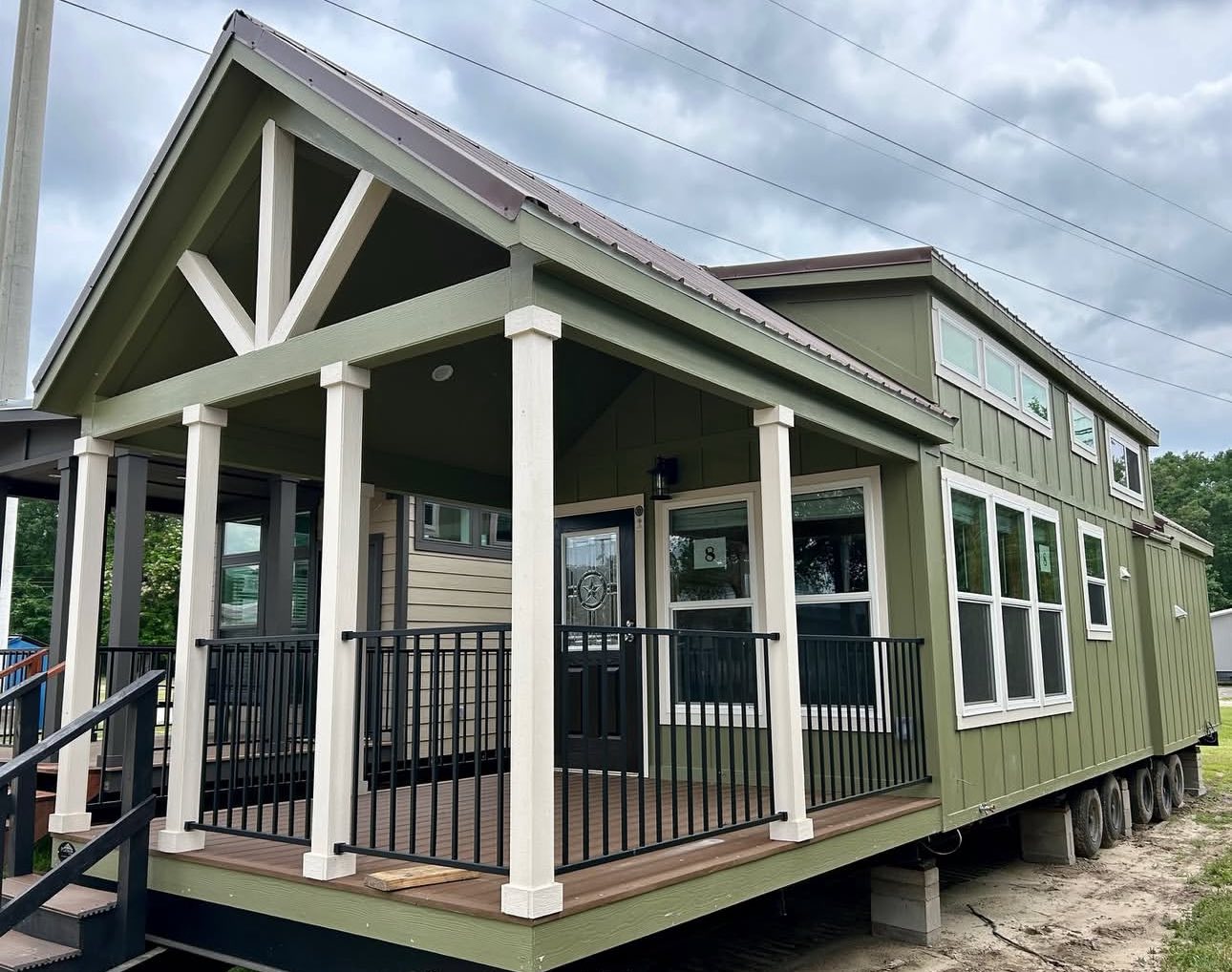 A green tiny house with a peaked roof and white trim sits on wheels. It features a covered porch with railings and large windows, under a cloudy sky.