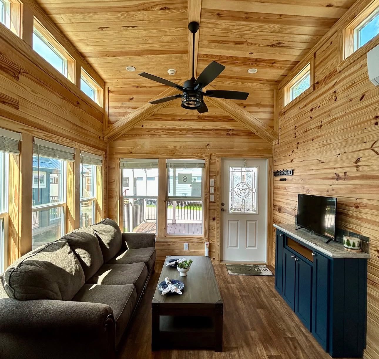 A cozy living room with wooden walls and ceiling, featuring a dark sofa, a ceiling fan, a wall-mounted TV on a cabinet, and large windows providing ample light.