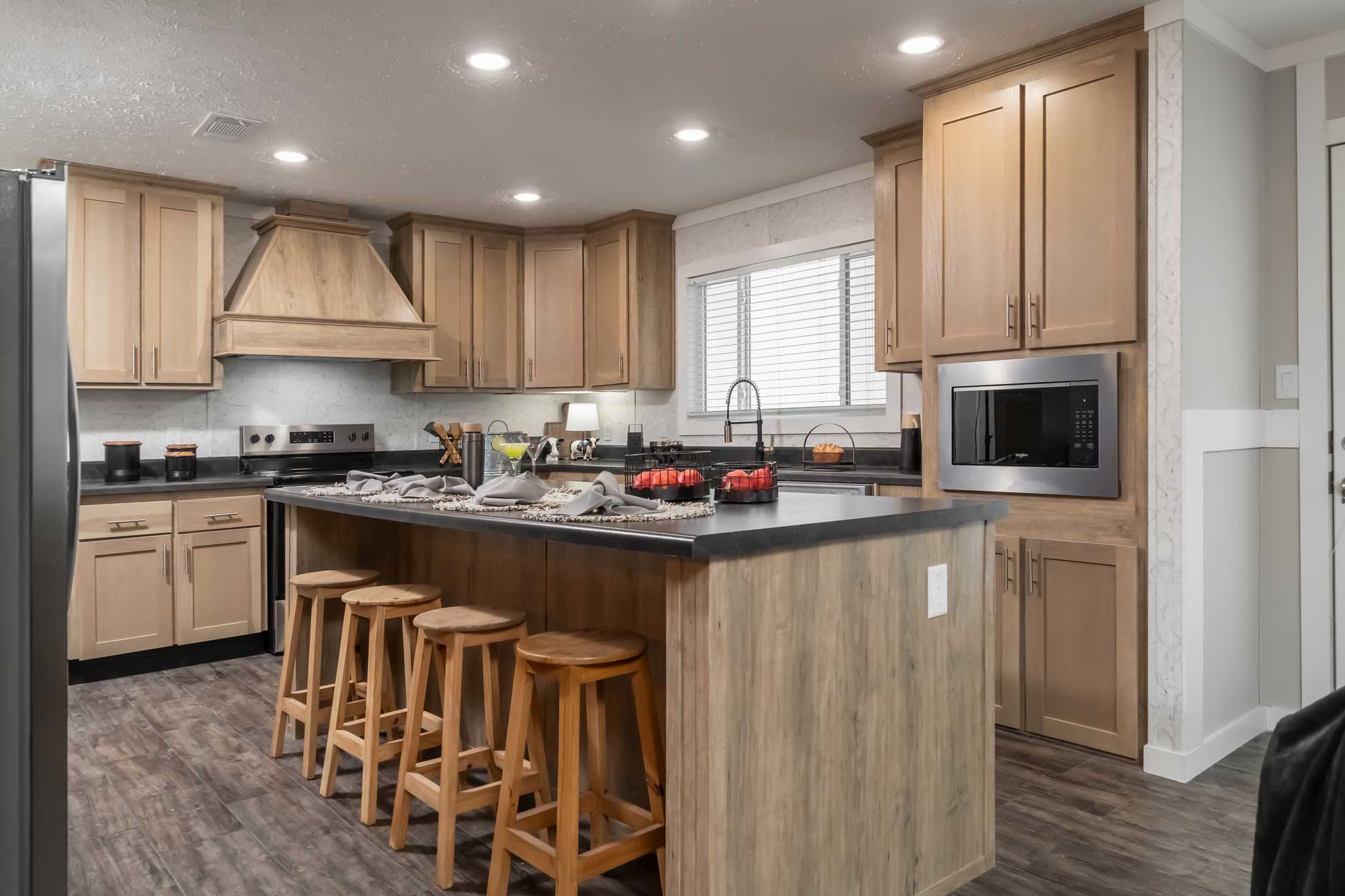 A modern kitchen with wooden cabinets and a large island. Four stools line the island, set with a gray cloth and decor. Soft lighting creates a cozy atmosphere.