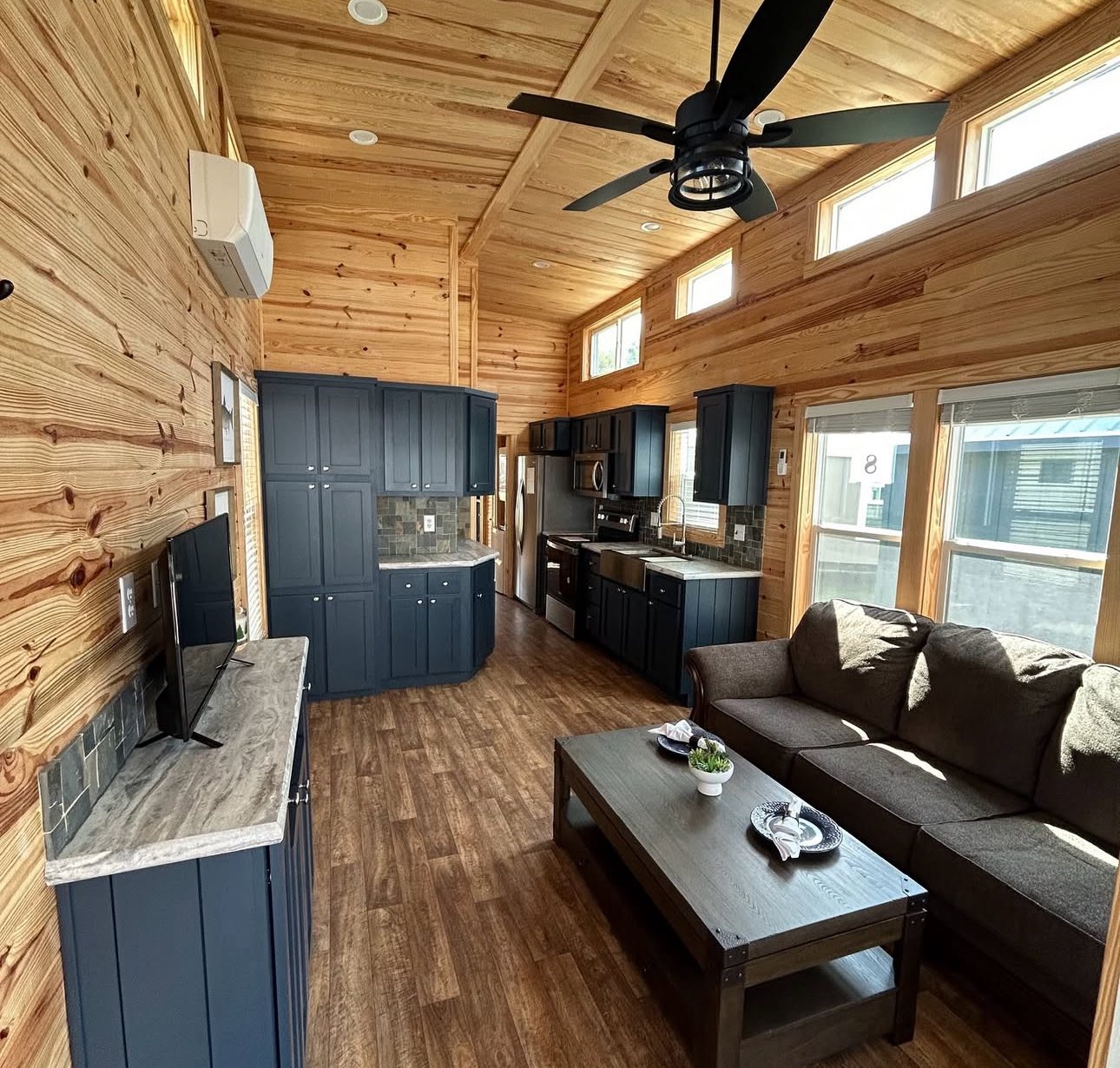 Rustic wooden interior featuring a cozy living area with a dark sofa, coffee table, and mounted TV. The open kitchen has navy cabinets and stainless steel appliances, under a ceiling fan and natural light from high windows.