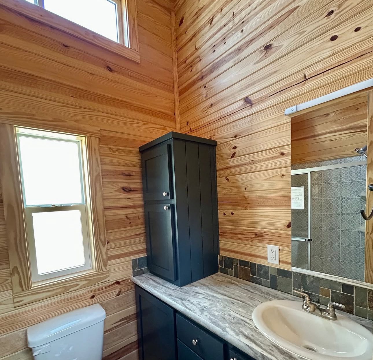 A cozy bathroom with natural wood paneling, a marble countertop, black cabinets, a white sink, and a window letting in soft natural light.