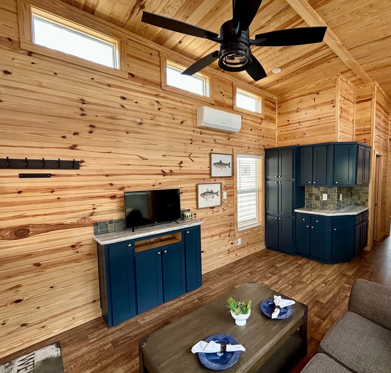 Cozy cabin interior with warm wood walls and ceiling, blue cabinets, a mounted TV, framed fish art, and a table set for two, creating a rustic vibe.