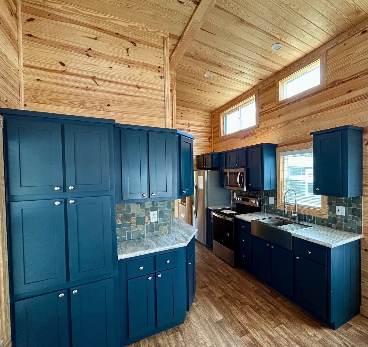 Modern kitchen with wood-paneled walls, blue cabinets, and stainless steel appliances. Natural light from windows enhances the cozy, rustic feel.