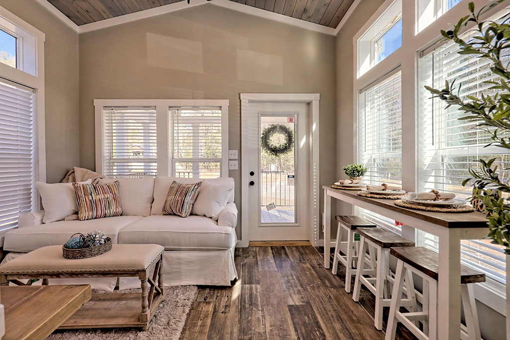 Bright living room with high ceilings, featuring a white sofa with striped cushions, wooden coffee table, and a door with a wreath. Sunlit space with serene ambiance.