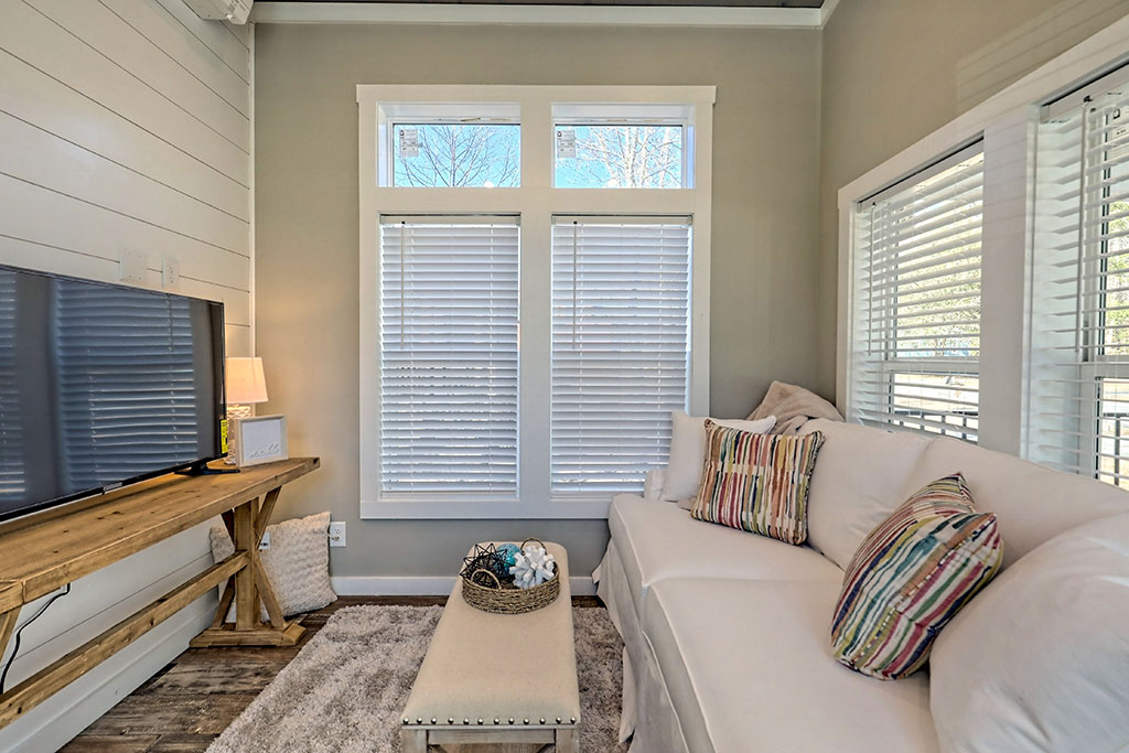 Cozy living room with a white sofa adorned with colorful striped pillows. A wooden table supports a TV, and large windows let in natural light.