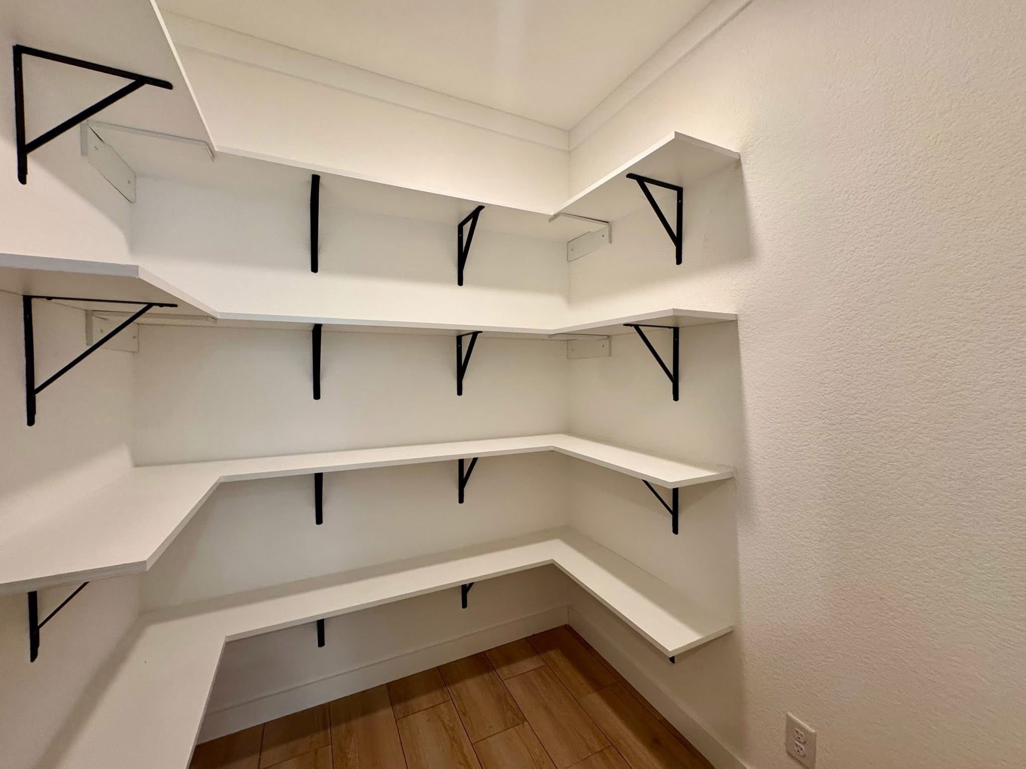 White pantry shelves with black brackets line the walls, forming an L-shape. The room has a warm wood floor and a minimalist, clean aesthetic.