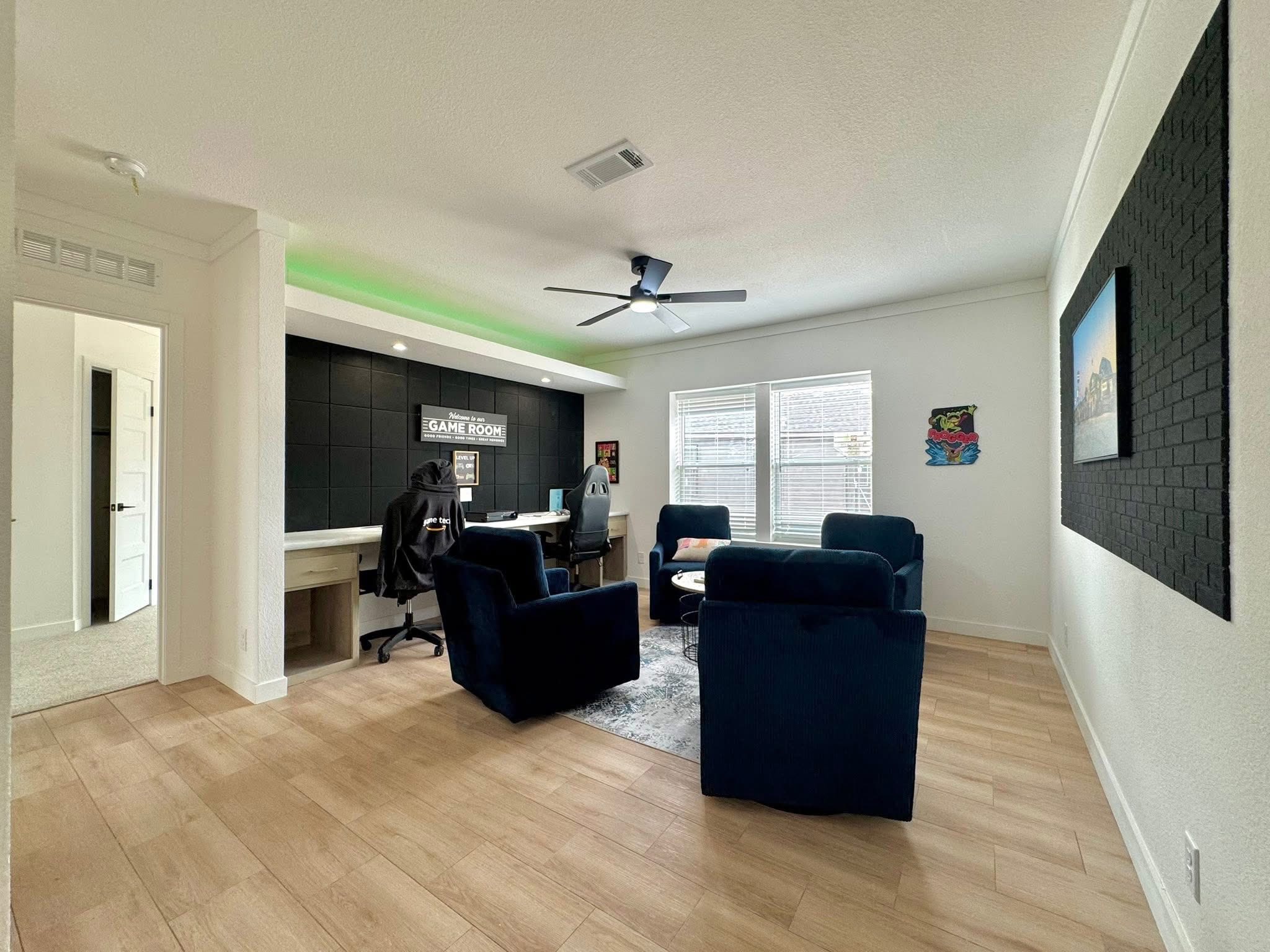 Modern game room with blue chairs, table, and gaming setup under a "Game Room" sign. Large windows, wood floor, and black accented wall.