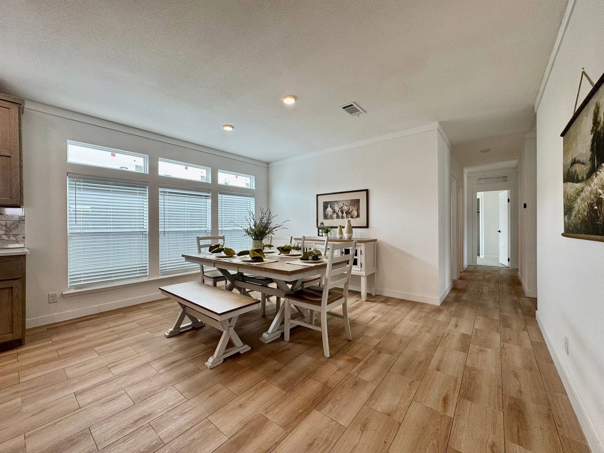 Bright dining area with a rustic table, benches, and chairs. Large windows illuminate the room. The decor includes greenery and framed art. Cozy and inviting atmosphere.