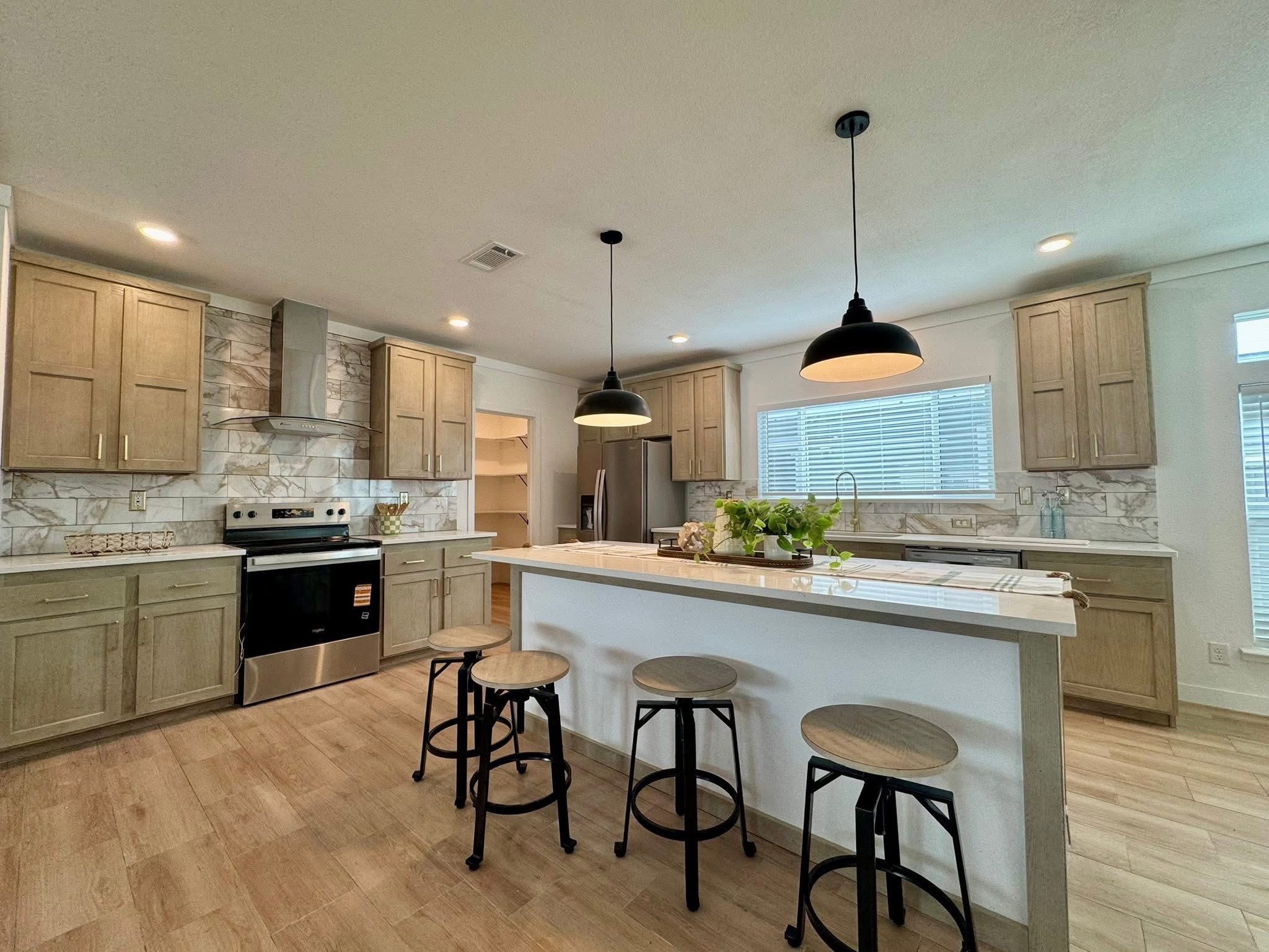 Modern kitchen with light wood cabinets, marble backsplash, and stainless steel appliances. An island with bar stools sits beneath pendant lighting.