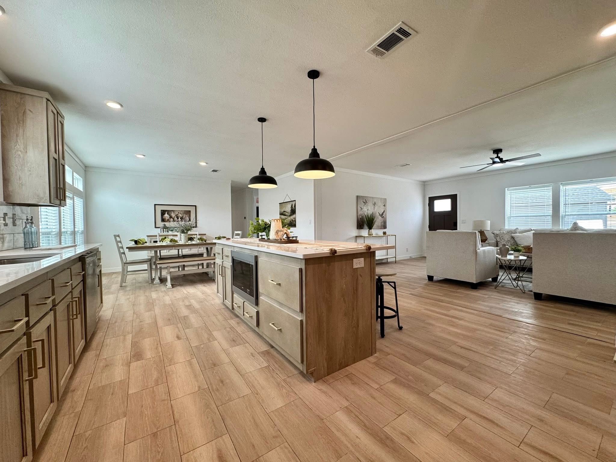 Spacious open-plan kitchen and living area with light wood flooring, modern pendant lights over an island, and cozy seating. Bright and inviting ambiance.