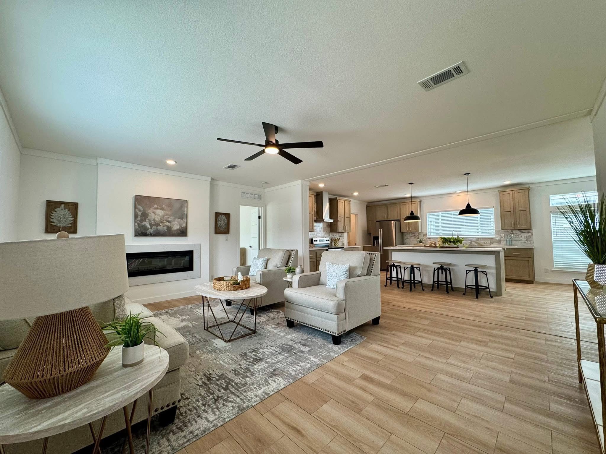 A spacious, modern living room and kitchen with neutral tones. Features comfy sofas, a large rug, ceiling fan, kitchen island with stools, and bright lighting.