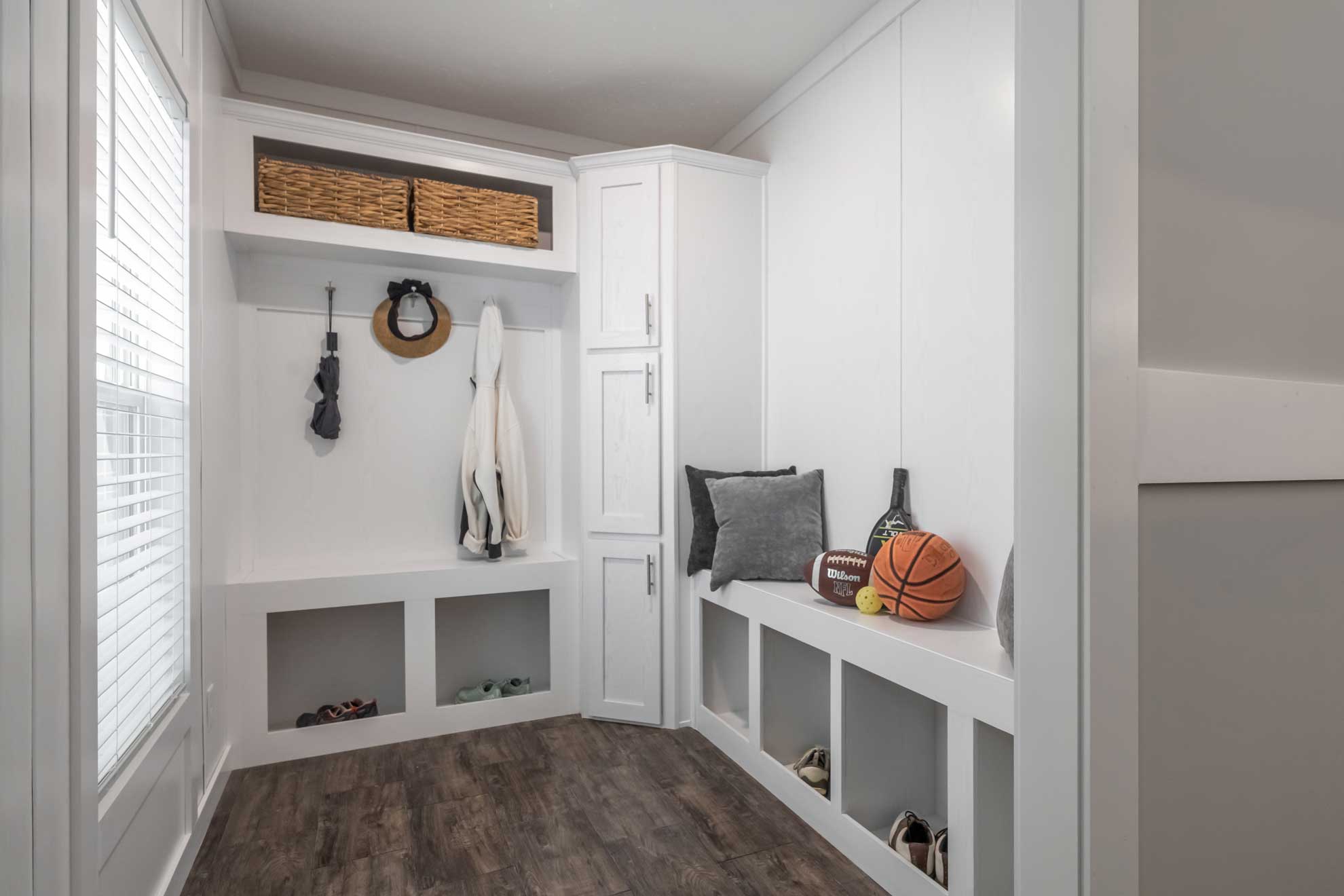 Modern mudroom with white cabinets and shelves on dark wood flooring. Hooks hold coats and hats, and baskets, a basketball, and other items are stored neatly.