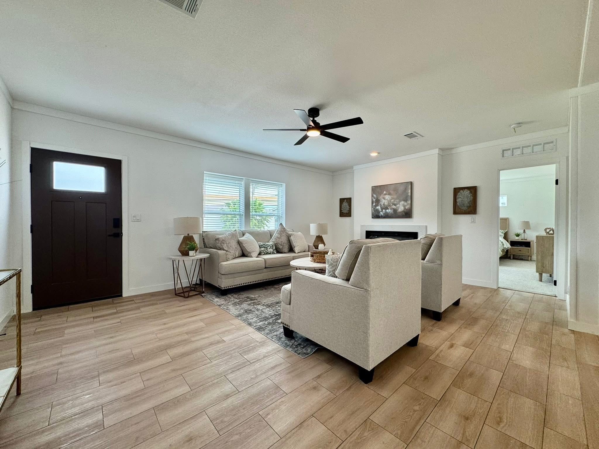 Spacious living room with light wood flooring, beige sofas, and armchairs. A ceiling fan hangs overhead. Soft lighting creates a cozy and inviting ambiance.
