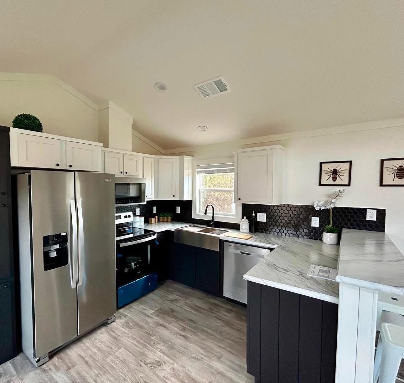 Modern kitchen with stainless steel appliances, white upper cabinets, black lower cabinets, hexagonal tile backsplash, and wood flooring. Cozy and chic.