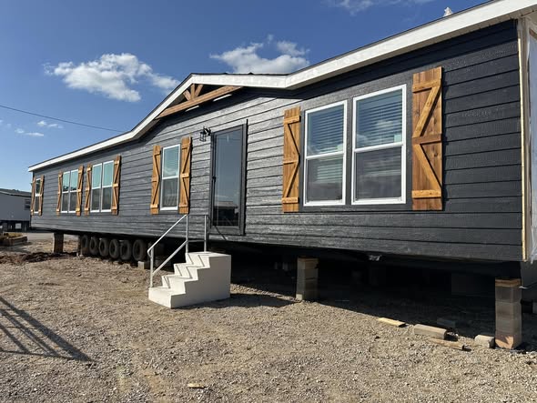 A dark gray manufactured home with rustic wooden shutters on multiple windows, set on a gravel lot under a blue sky, conveys a modern, cozy feel.