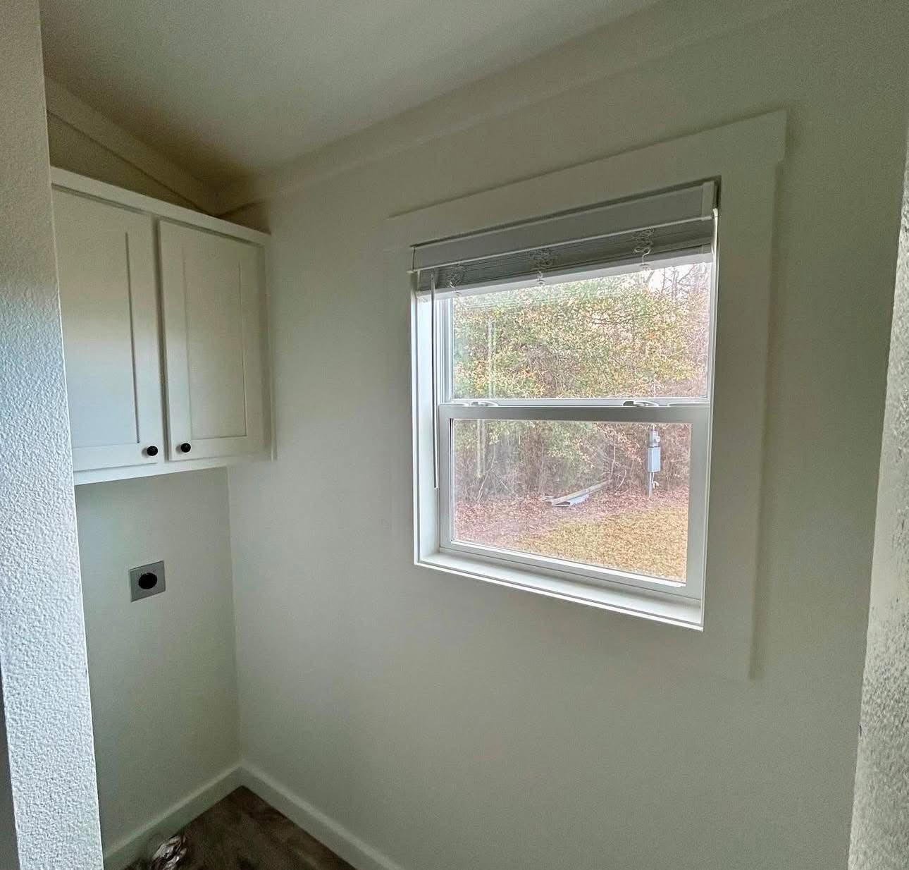 A small, white laundry room with a window overlooking a garden. The room features white cabinets, a blank wall, and a cutout space for a dryer.
