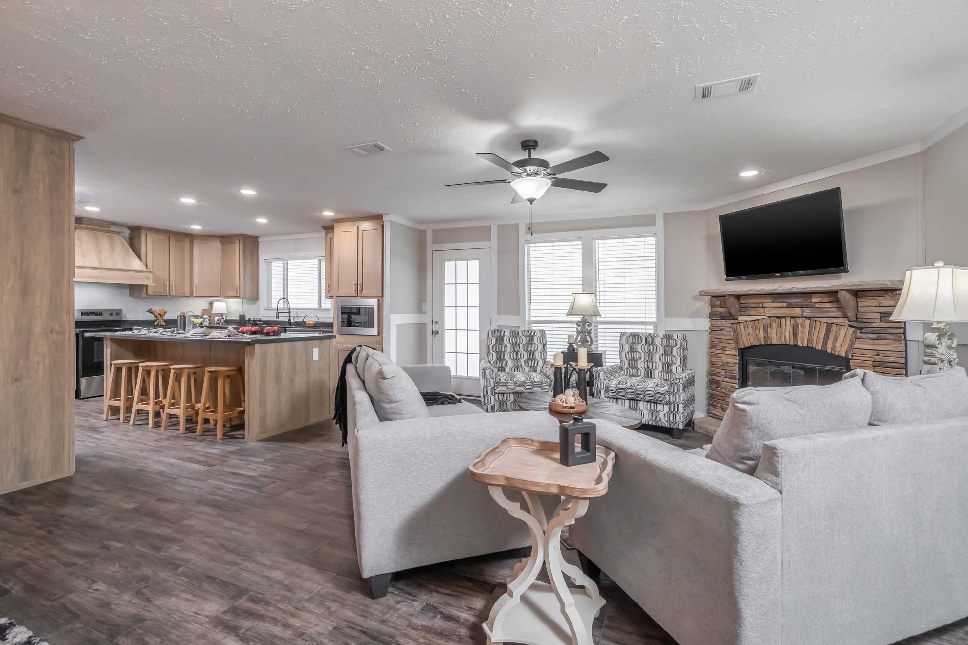 Spacious living room with gray sofas, stone fireplace, and wall-mounted TV. Open kitchen features wood cabinets and island with stools. Cozy ambiance.