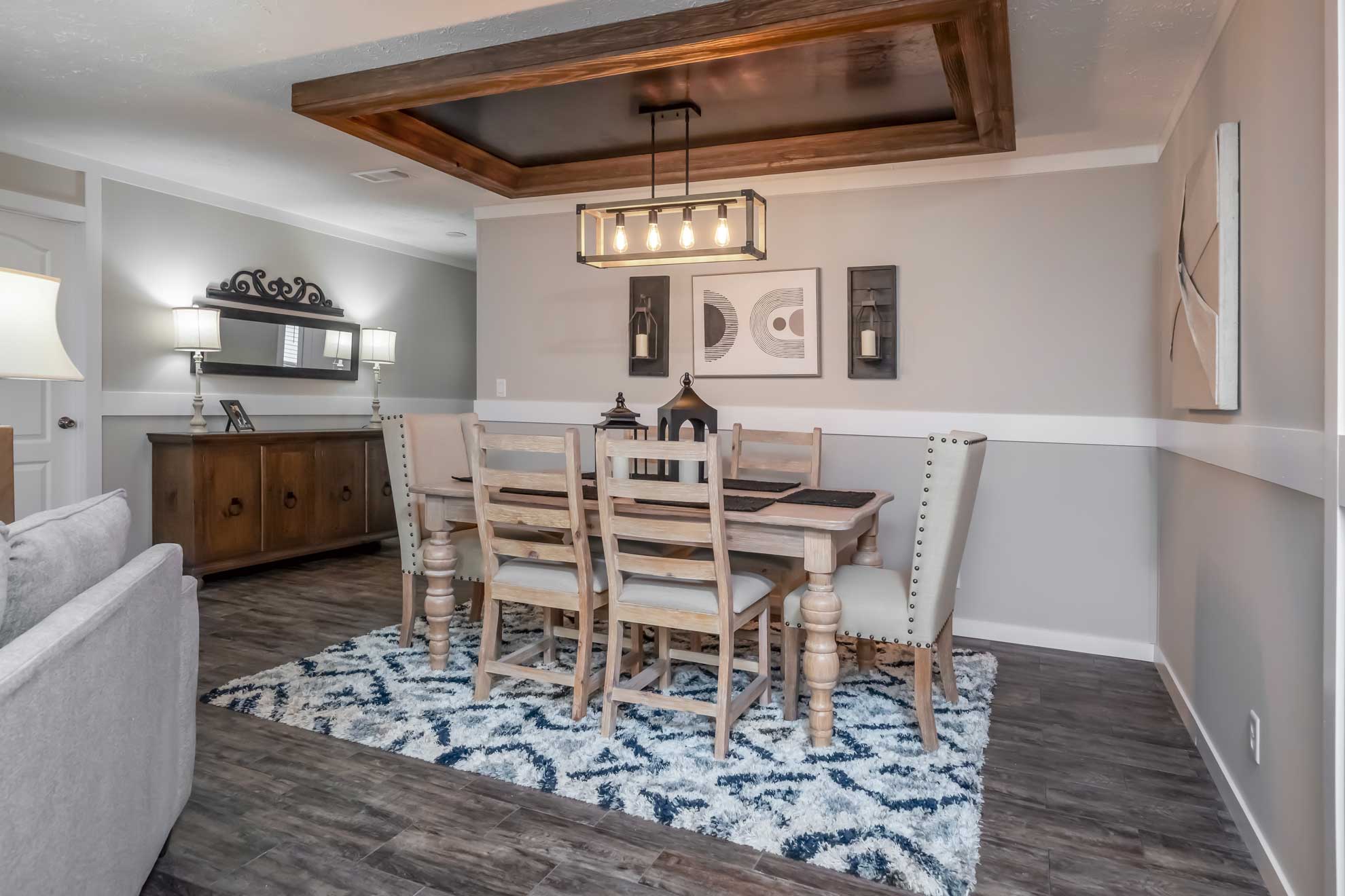 Dining room with a wooden table and six chairs on a patterned rug. Modern chandelier, abstract wall art, and a sideboard with lamps and a mirror enhance the cozy ambiance.