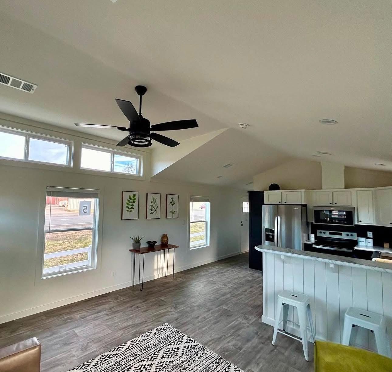 Modern open-concept living area with vaulted ceilings and natural light. Kitchen features stainless steel appliances; wall art and green stools add charm.