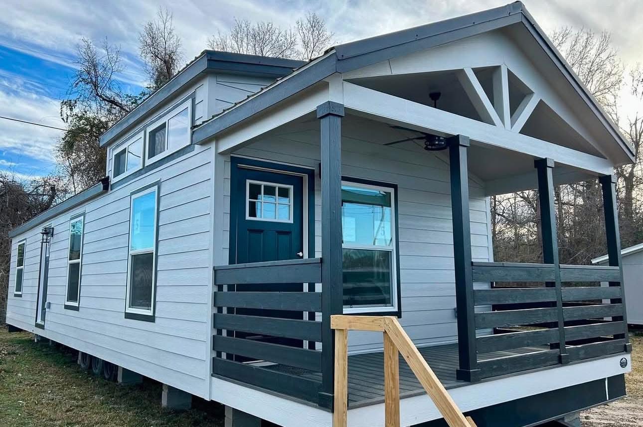 A modern tiny house with grey siding, a dark green door, and a covered porch with railing. It sits on grassy ground under a blue sky with clouds.