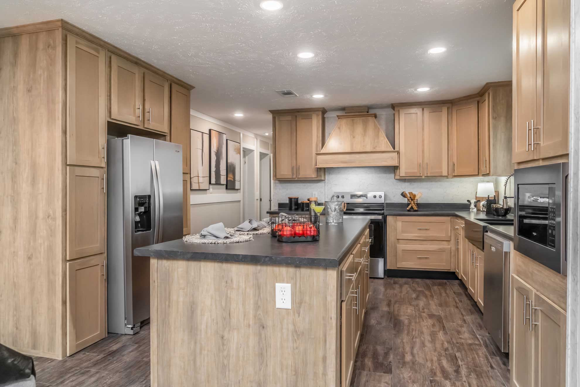 Modern kitchen with light wood cabinets, dark countertops, and stainless steel appliances. An island with red apples and decor provides a cozy, warm feel.