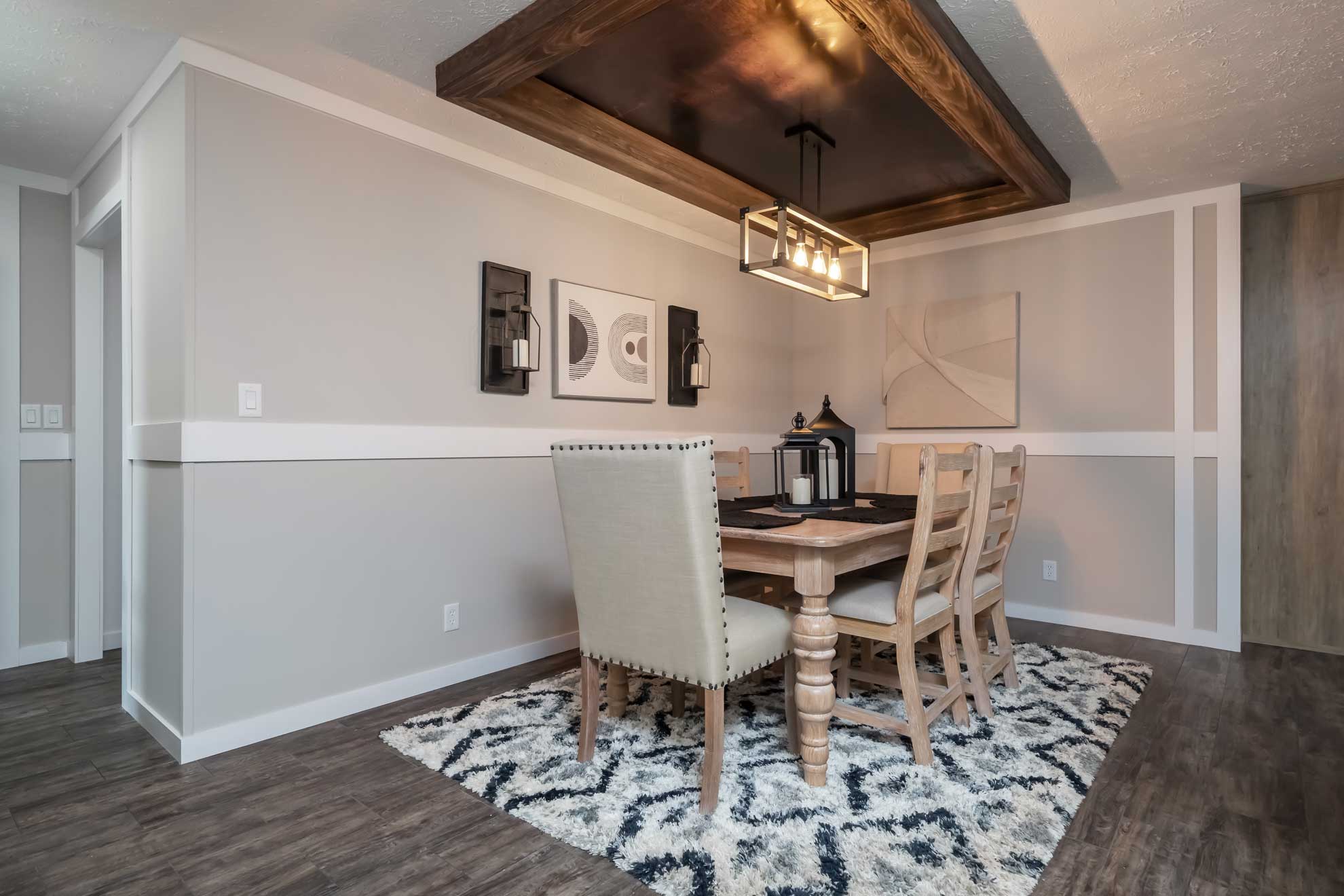 Modern dining room with a wooden table and six chairs on a patterned rug. Stylish ceiling light, abstract wall art, and neutral tones create a cozy atmosphere.