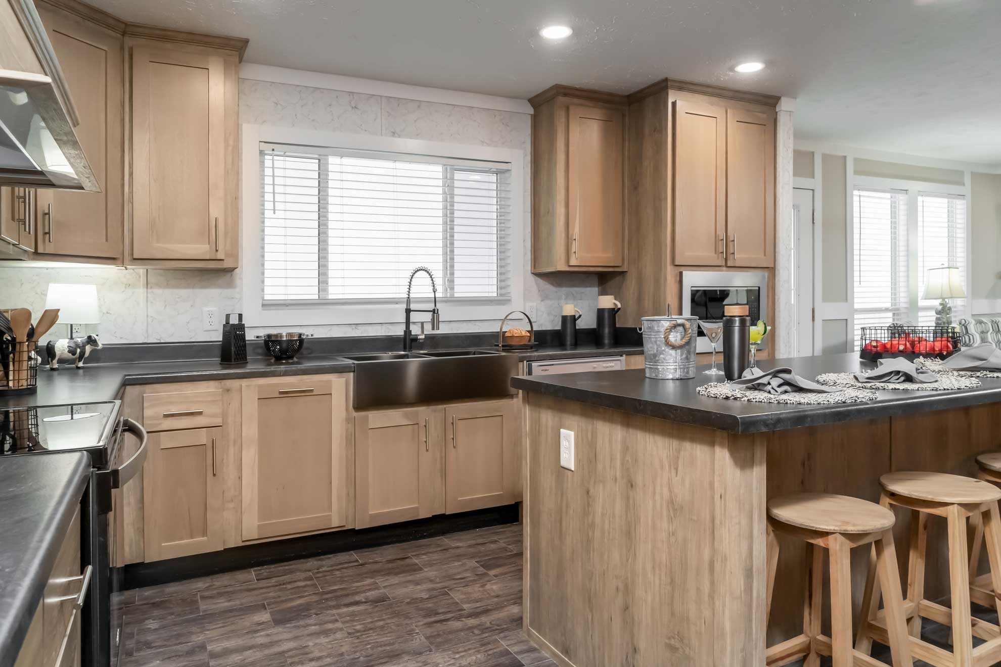 Modern kitchen with light wood cabinets, dark countertops, and a large island with wooden stools. Stainless steel appliances and a farmhouse sink add a sleek touch.
