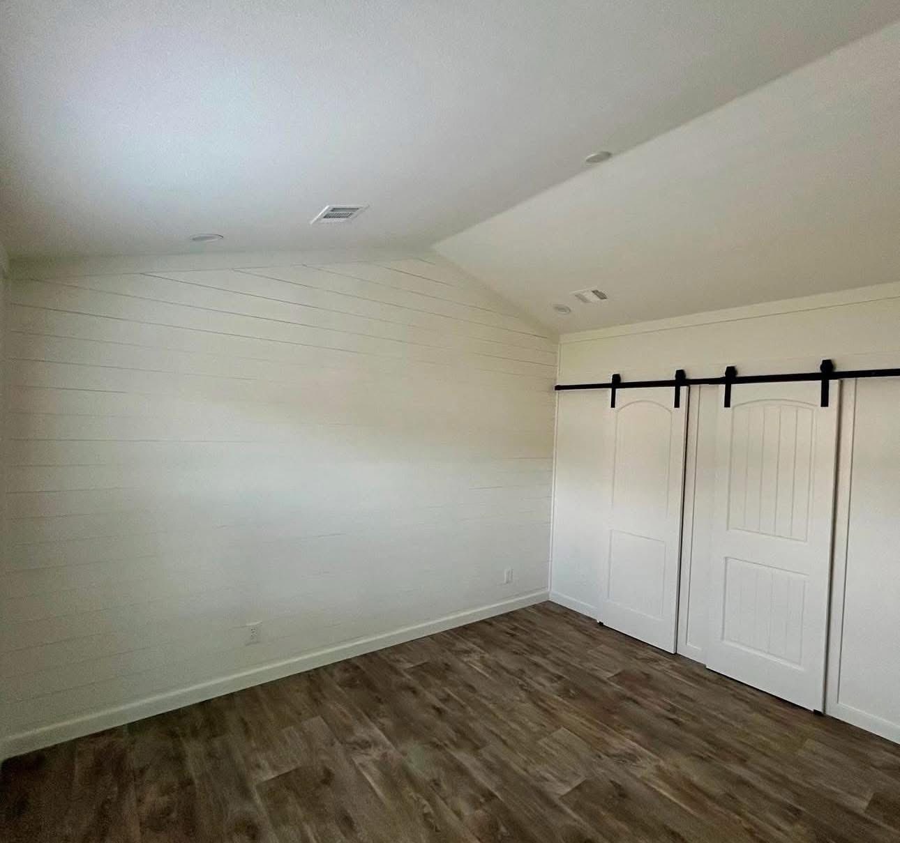 An empty room with white shiplap walls and a vaulted ceiling. The floor is dark wood, and there are white sliding barn doors on the right. Bright and clean atmosphere.