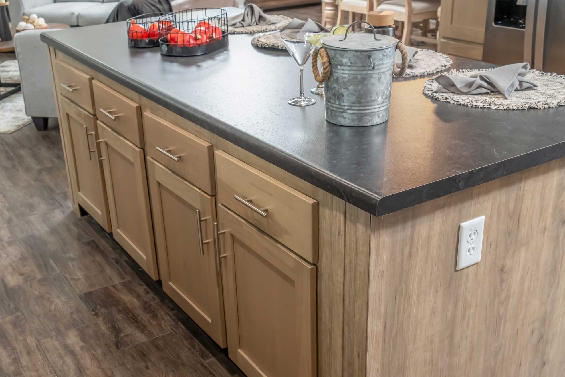 Spacious kitchen with a black countertop island, light wood cabinets, a wire basket with tomatoes, a tin bucket, and two empty martini glasses.