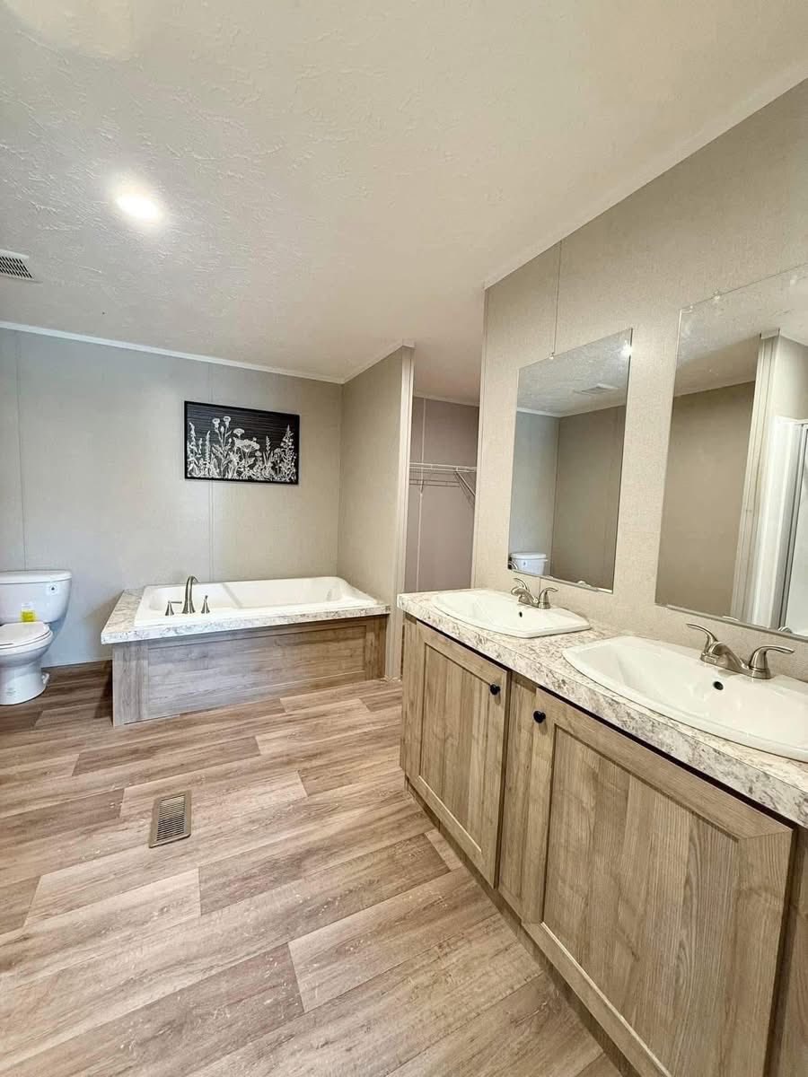 Spacious bathroom with dual sinks on a wooden vanity, a large tub, toilet, and a monochrome floral artwork on the wall. Light wood flooring.