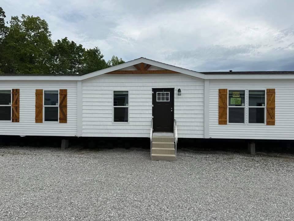 Single-story white mobile home with a brown door and steps. Wooden shutters frame three windows. Gravel driveway and trees in the background. Overcast sky.