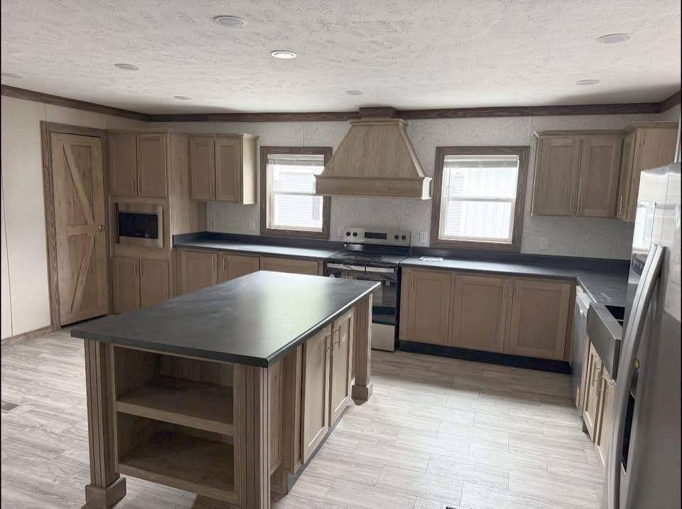 Spacious kitchen with wooden cabinets, central island, dark countertops, and stainless steel appliances. Natural light from two windows, calming tone.