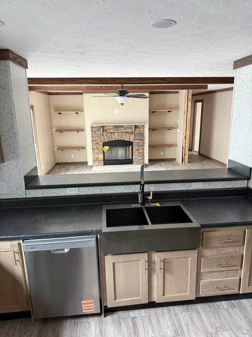Modern kitchen overlooking a cozy living room with a stone fireplace. Stainless steel dishwasher and black countertop add a sleek touch.