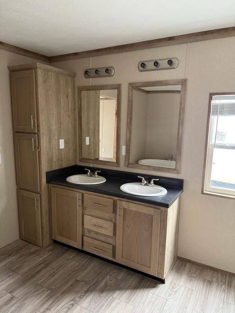 Modern bathroom with dual sinks on a dark countertop, wooden cabinets, two mirrors above, and a tall storage cabinet on the left. Natural light from a window.