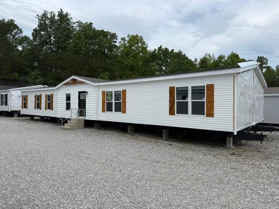 A single-story white mobile home with wooden shutters is elevated on risers in a gravel area. Trees are visible in the background under a cloudy sky.