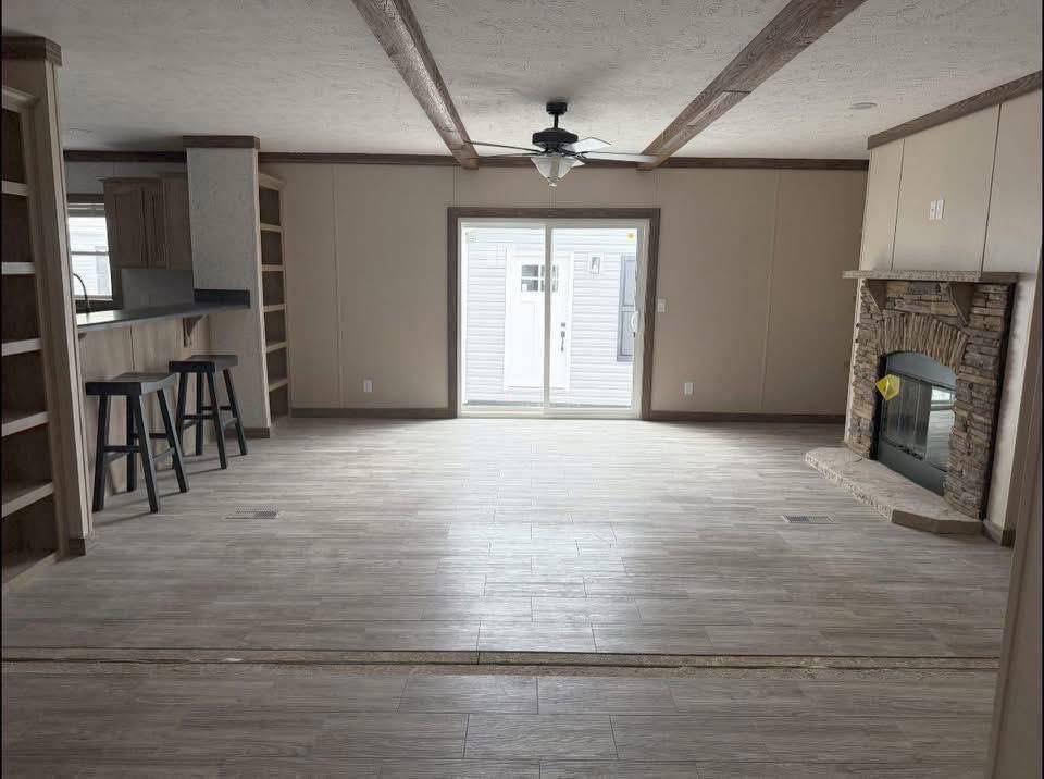 Spacious living room with light wood flooring, a ceiling fan, and a stone fireplace on the right. A breakfast bar and stools are on the left.