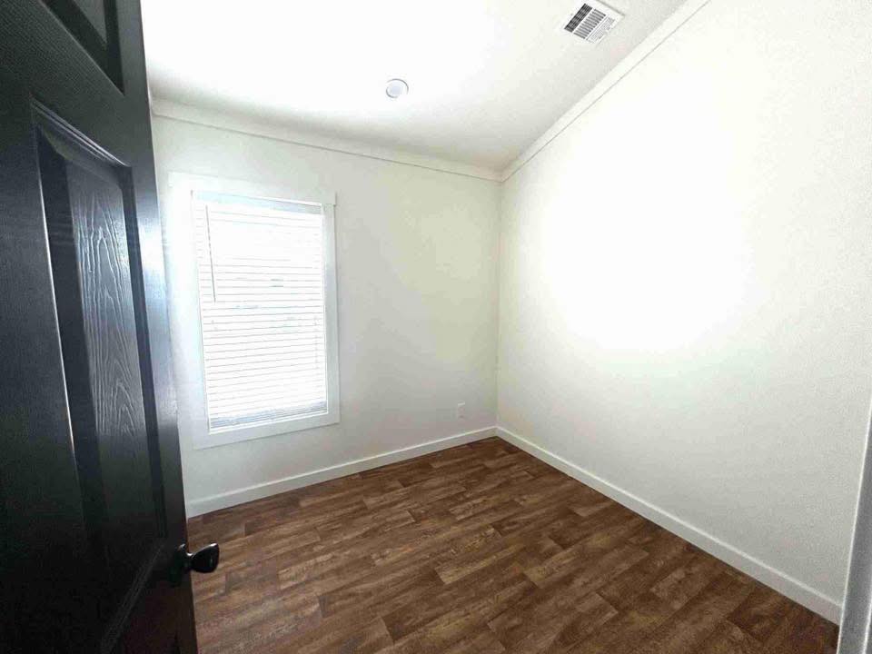 Small, empty room with white walls and a wood-paneled floor. A single window with blinds lets in light, and a black door is partially open.