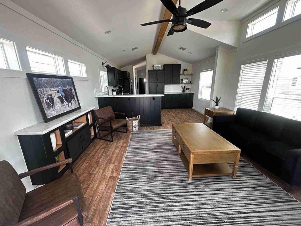 Modern living room with wooden floor and ceiling beam, featuring a black sofa, striped rug, and a wooden coffee table. Bright and airy atmosphere.