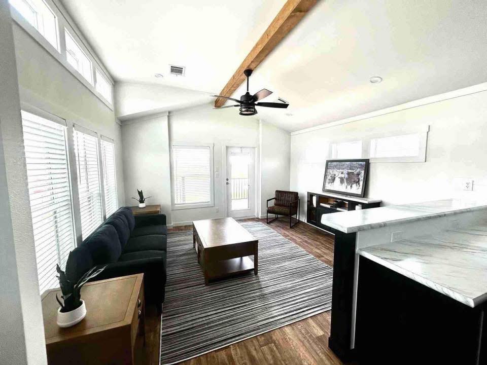 Modern living room with high ceiling, wooden beam, and ceiling fan. Features a dark sofa, striped rug, and wall art above a console table. Bright, airy ambiance.