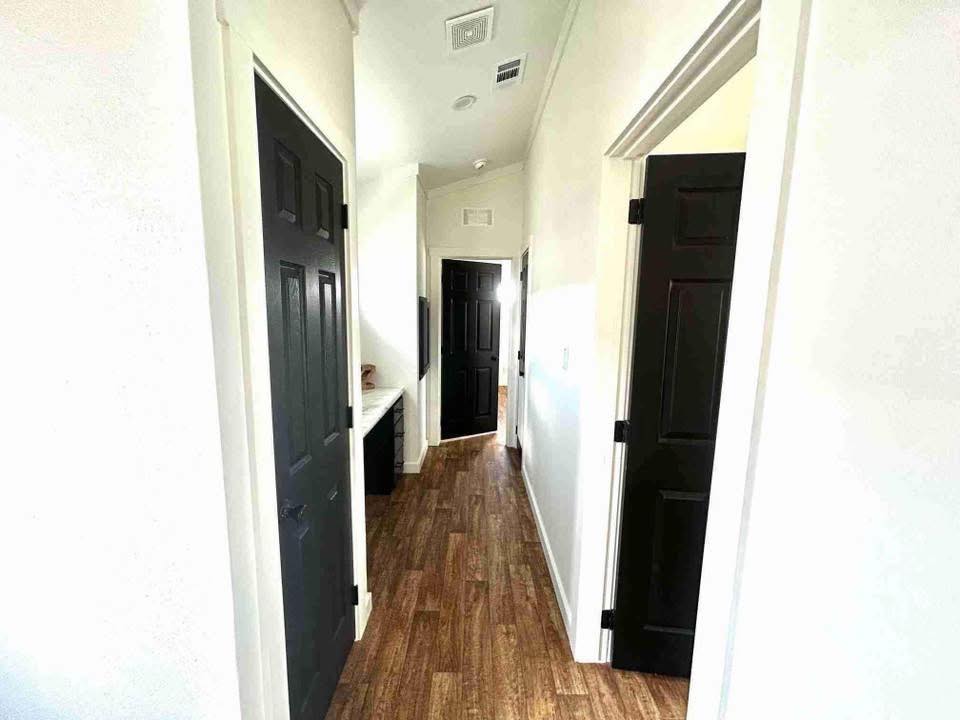 A narrow hallway with light wood flooring and white walls. Several black doors line the corridor, with bright lighting casting a clean, modern feel.